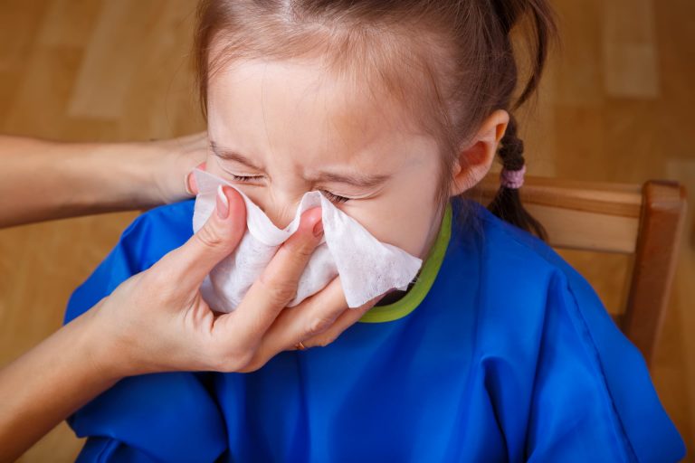 Stock photo of a kid sneezing while someone holds a tissue over their nose