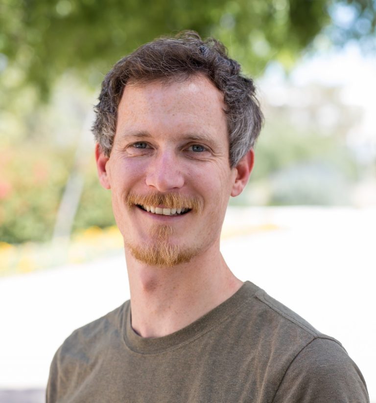 Tom Riffelmacher, Ph.D., looks into the camera. He is standing outside and smiling