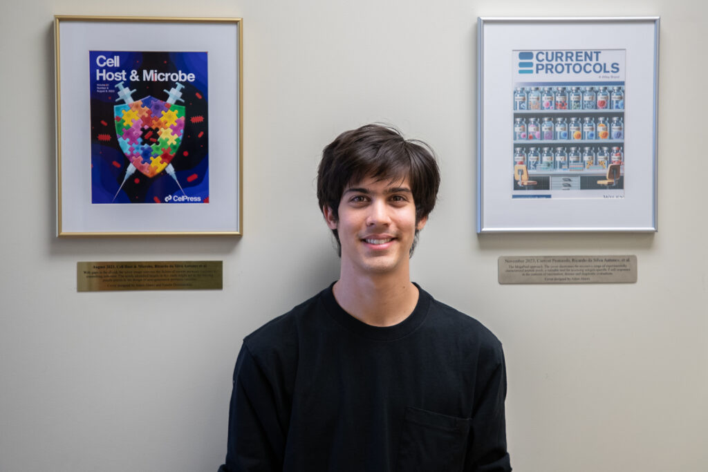 Photo of Adam Abawi posed between two framed journal covers. The cover on the left is for Cell Host and Microbe. The cover on the right is for Current Protocols.