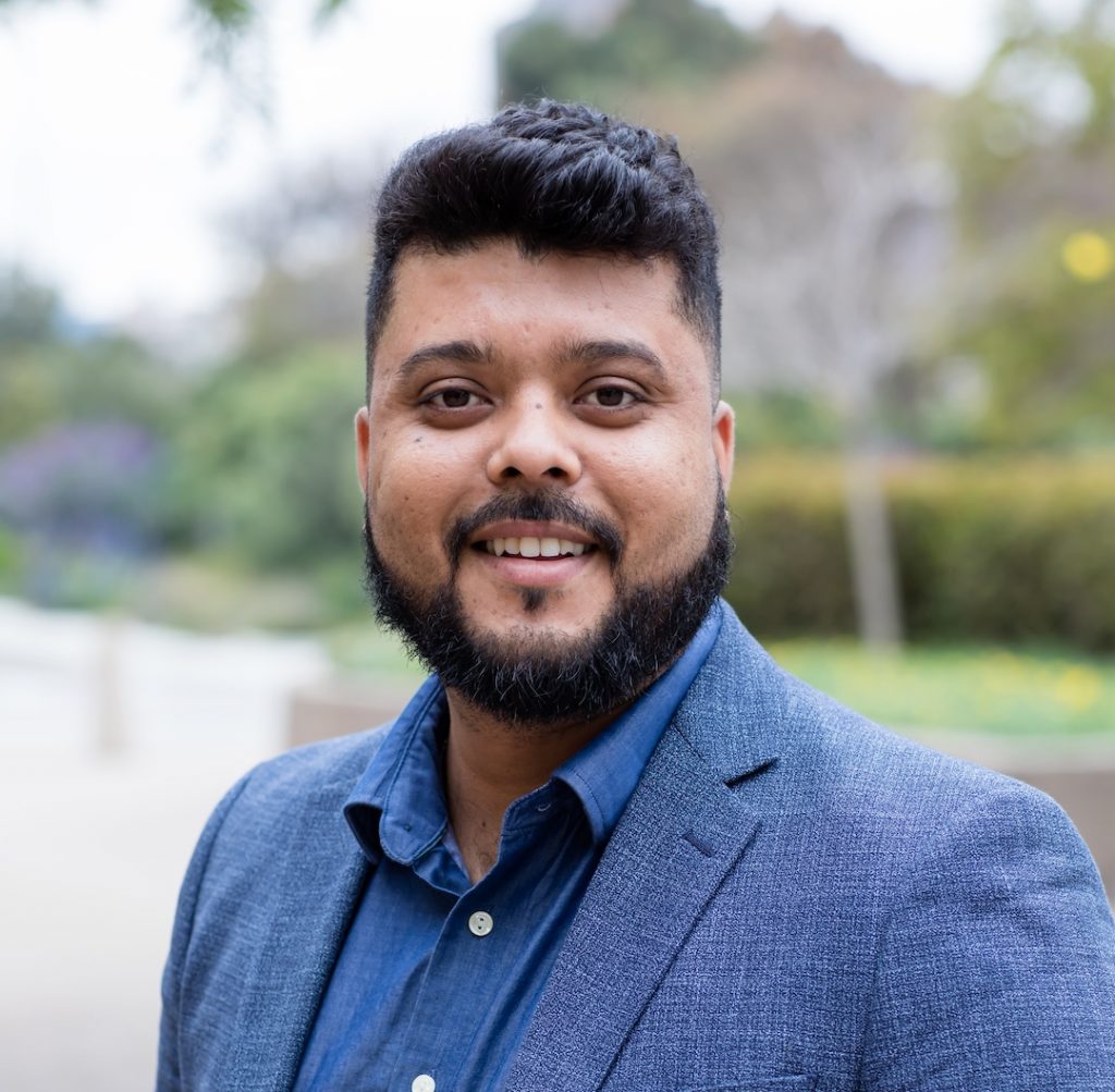 Portrait photo of Alves. He is standing outside in a blue suit and smiling