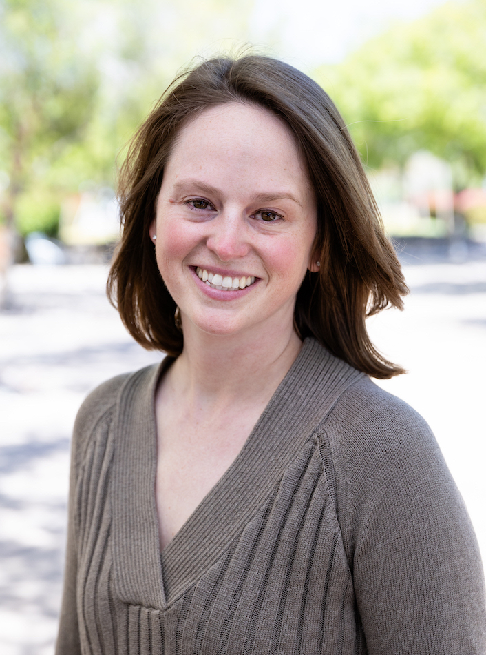 Scientist Rachel Helms poses for the camera. She is outside. She is smiling. She has brown hair and is wearing a brown top.