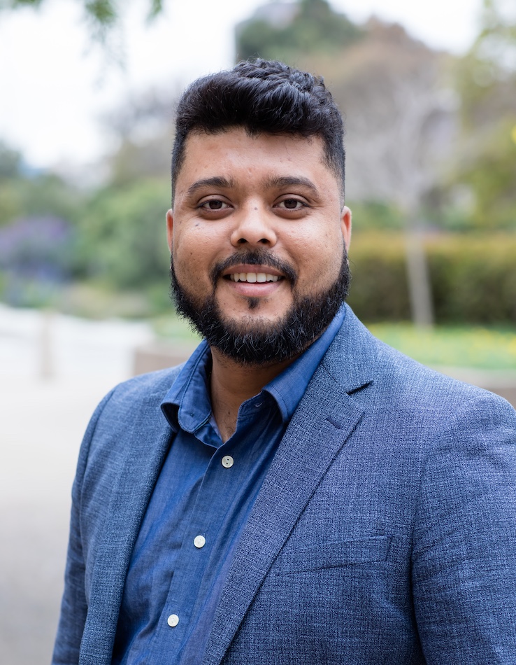 Rubens Alves smiles at the camera. He is has dark hair and a dark beard. He is wearing a blue collared shirt and blazer. 