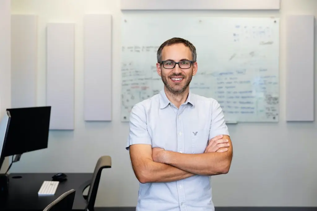 Photo portrait of LJI Research Assistant Professor Benjamin J. Schmiedel, Ph.D. He is standing in an office setting and smiling at the camera