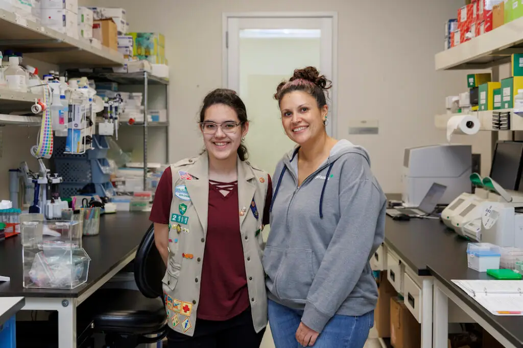 Alarcón and Brielle in a lab setting. Brielle is wearing a khaki Girl Scout vest with patches and badges