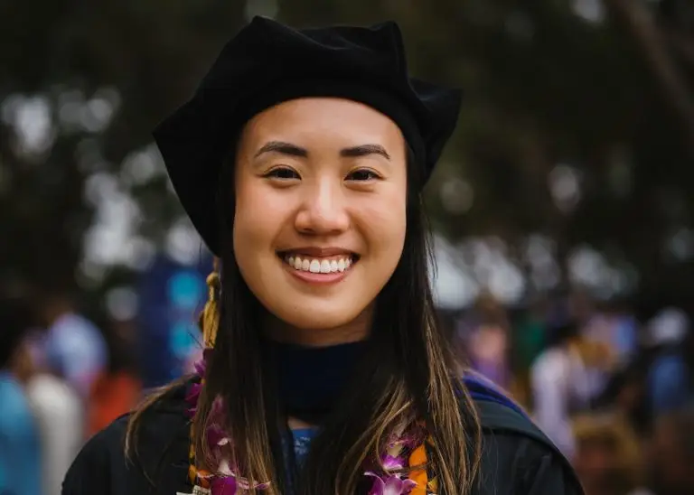 Ivy Phung faces the camera and smiles. She is wearing a black graduation robe and cap. She is wearing a necklace of flowers and a necklace of dollar bills.