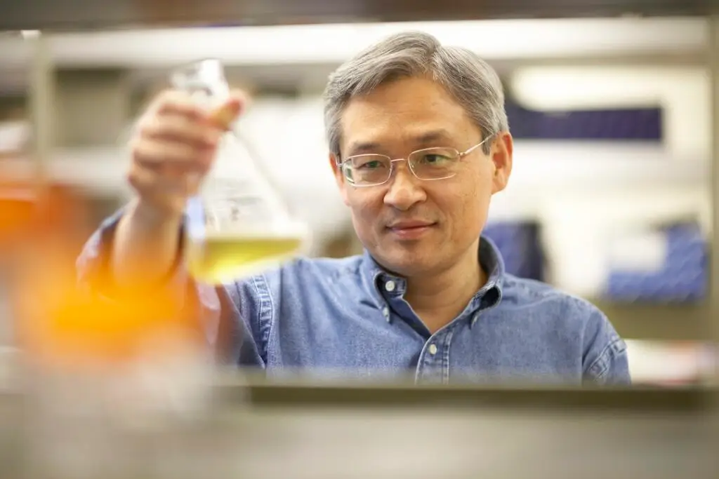 Photo portrait of Toshi Kawakami. He is posing, holding a flask in a lab setting