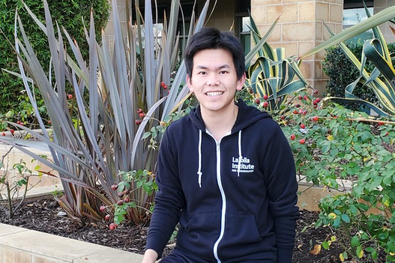 Wang sits on a small wall outside LJI. He is smiling at the camera