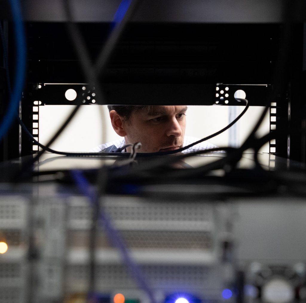 Michael Scarpelli is shown looking through a computer server area. His face is framed by machinery and wires.