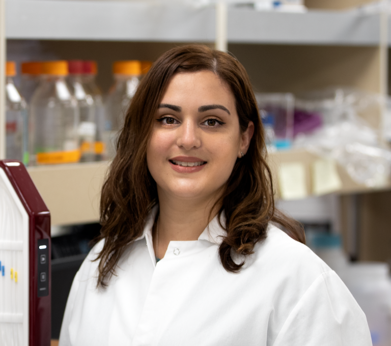 Ana-Sosa-Batiz stands in a laboratory. She is wearing a white laboratory coat and smiling at the camera