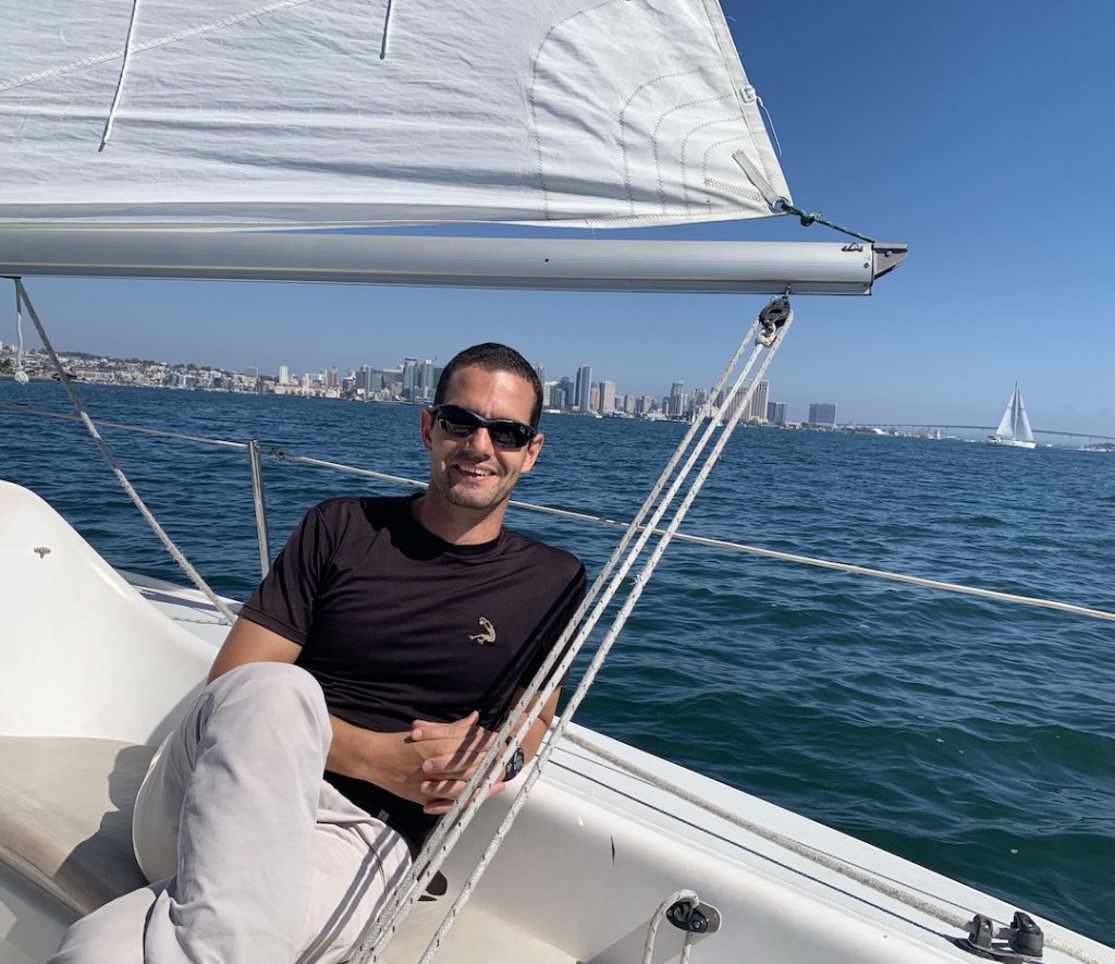 Marki sits on a sail boat. He is smiling and wearing sunglasses. The bay and San Diego skyline are behind him