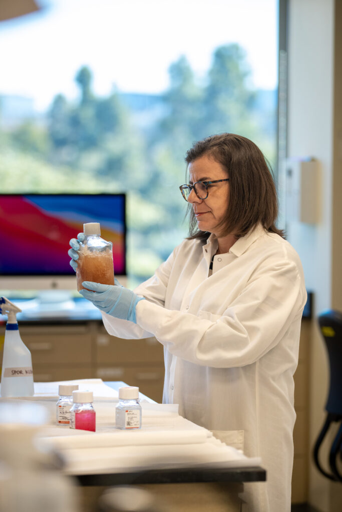 Bianca Mothé, Ph.D., in a lab setting. Wearing a white coat and holding a sample in a flask