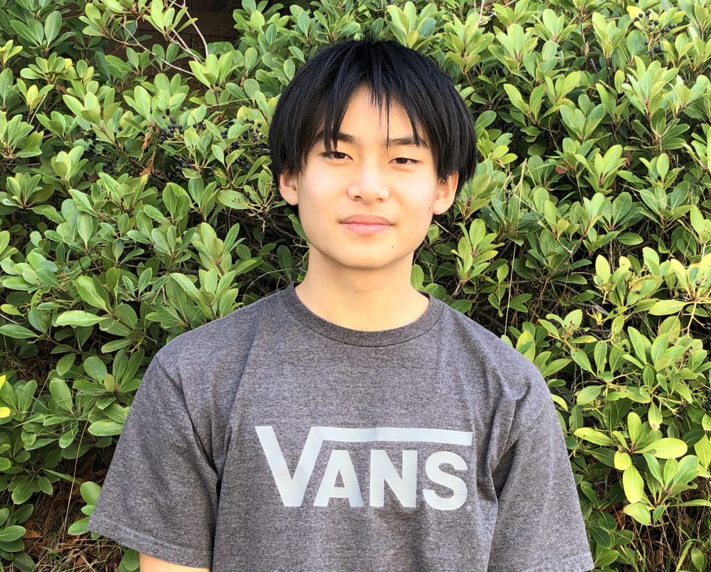 Photo of Daniel Cai. He is standing outside with leaves as his background. He is wearing a grey shirt