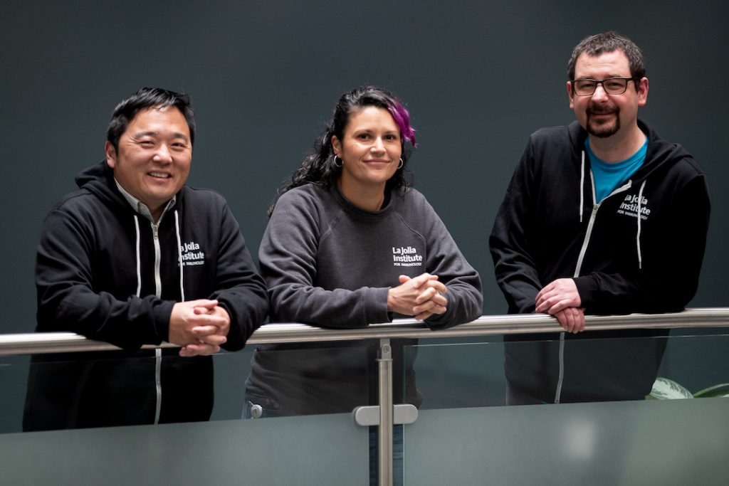 Three scientists stand on a balcony at LJI. Left to right: Kenneth Kim, Suzie Alarcon, and Zbigniew Mikulski