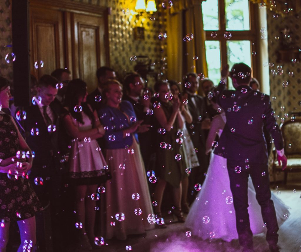 Photo of a crowd at a wedding. Bride is dancing. Bubbles fill the air. The lighting is purple.