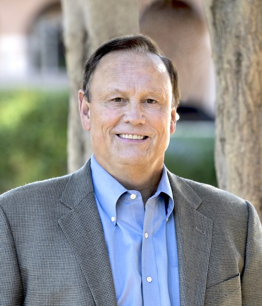 Portrait photo of David Rickey. He is wearing a grey suit and smiling at the camera