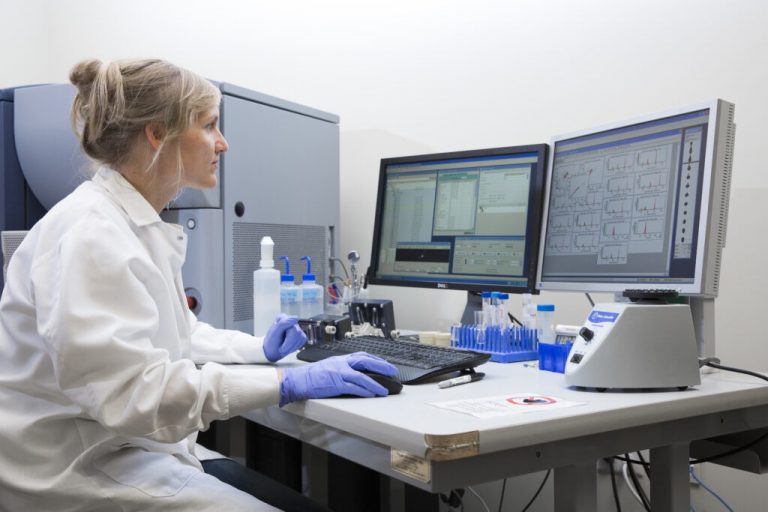 Photo of Denise Hinz working in the LJI Flow Cytometry Core. She is wearing gloves and a lab coat and looking at a computer