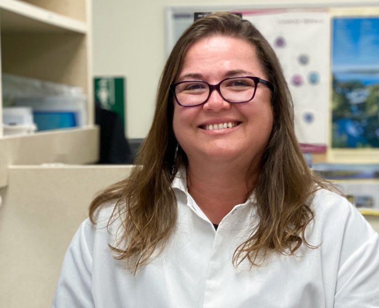 Roberta stands in a lab space. She is wearing a white lab coat and smiling