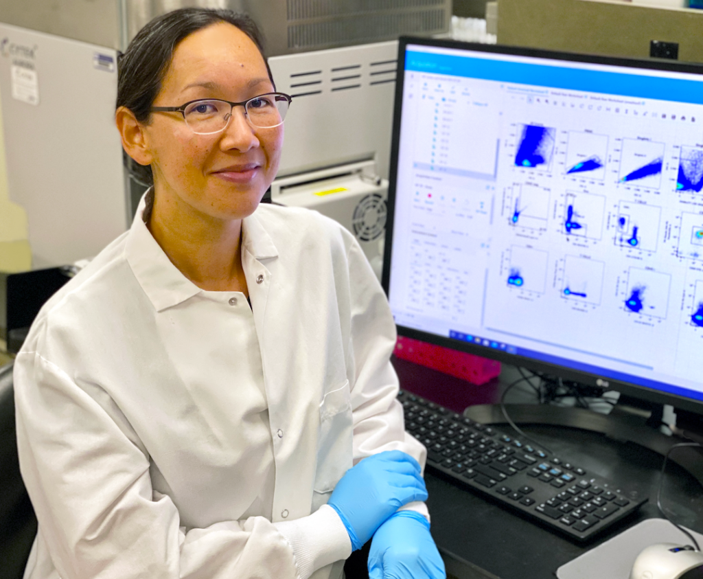 Ramirez sits at a computer in a lab space. She is wearing a white lab coat and looking at the camera