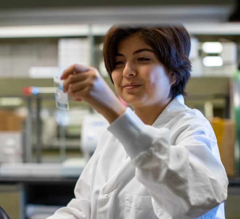 Intern Isabella Montoya in the laboratory. She is wearing a white lab coat and holding up a vial.