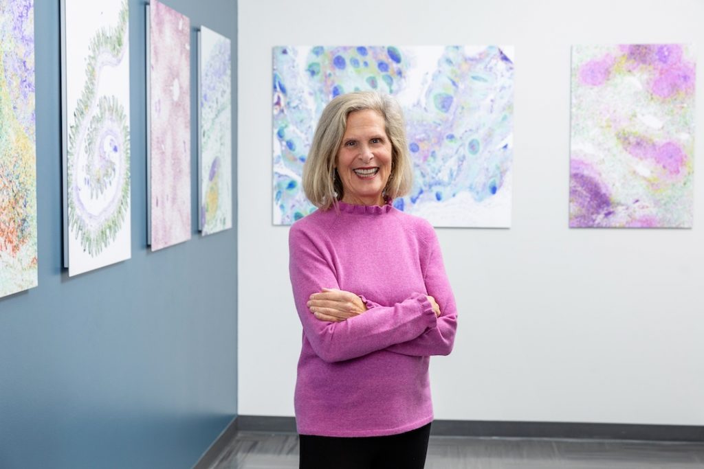 Barbara Donnell stands in front of mounted microscopy images in the LJI art gallery. Donnell is wearing a pink sweater and smiling at the camera